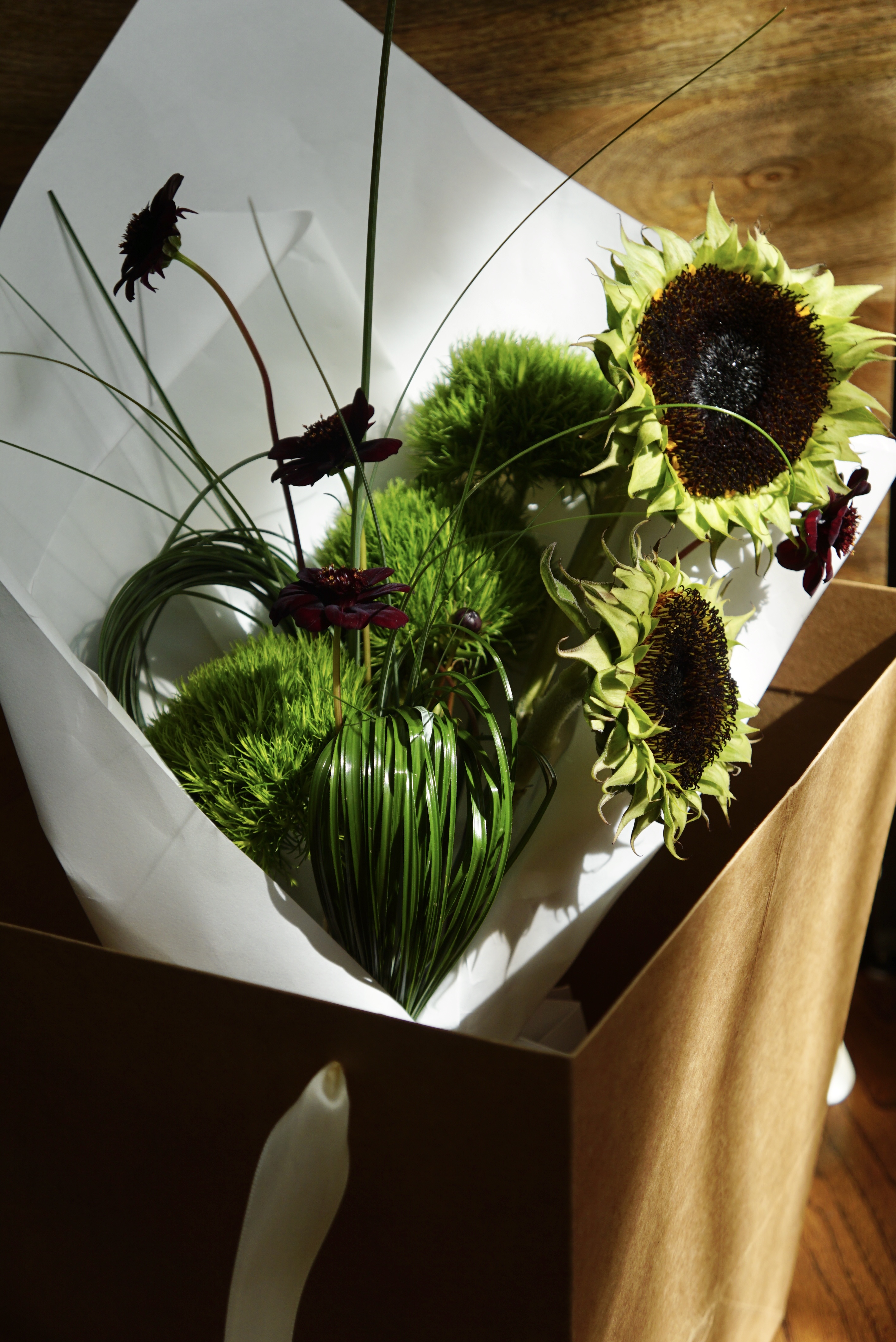 A modern flower bouquet with grasses, dianthus, chocolate cosmos, and naked sunflowers wrapped in white wrapping and sitting in a brown flower bag.