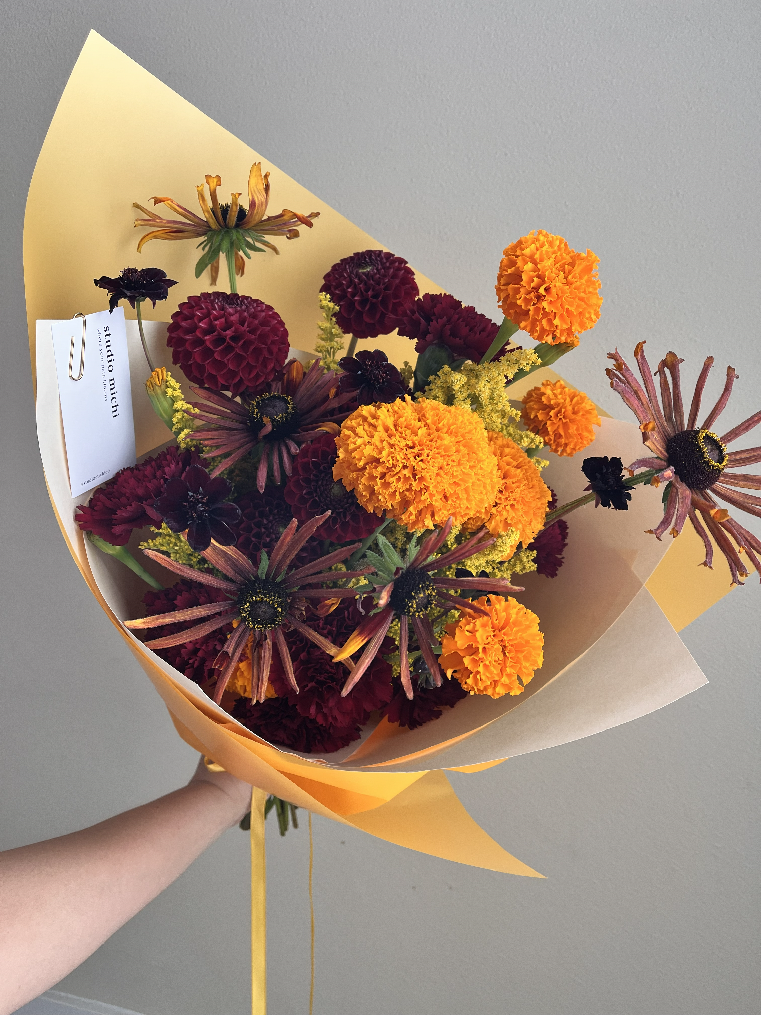 A hand-tied classic bouquet with warm-toned summer blooms wrapped in orange and cream paper with an orange ribbon. Seasonal flowers featured include orange marigolds, deep red dahlias, chocolate cosmos, orange and red rudbeckia, yellow solidago, and deep red carnations.