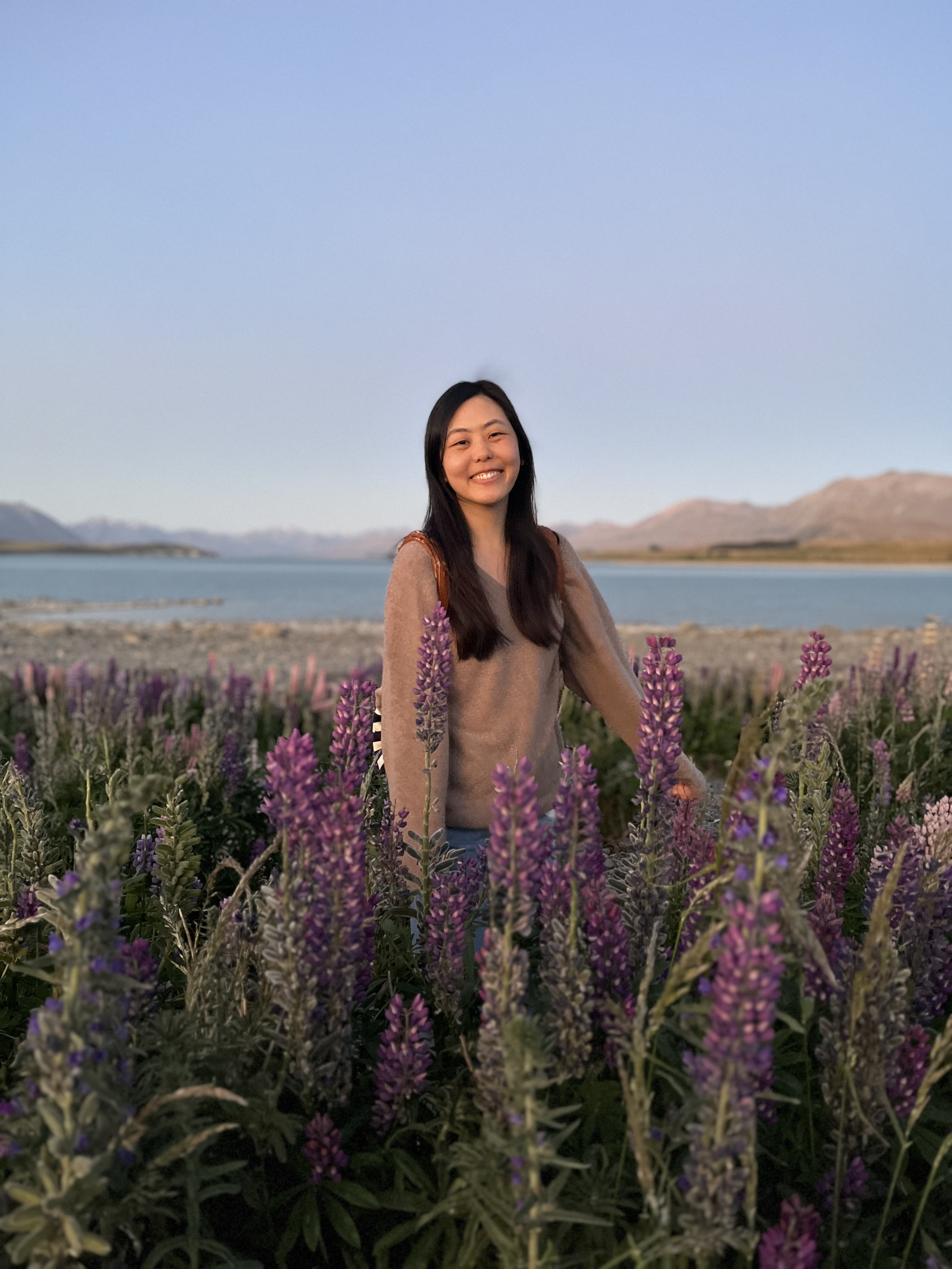 Jenn, the florist and owner of Studio Michi, smiling and standing in front of wild purple lupines with a view of a lake and mountains in the background.